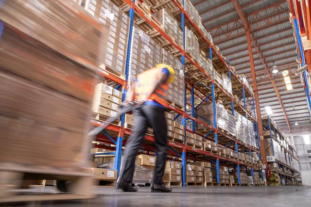 Worker in safety vest pulls a loaded pallet jack down an aisle of a high-rack warehouse