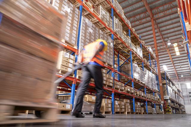 Worker in safety vest pulls a loaded pallet jack down an aisle of a high-rack warehouse