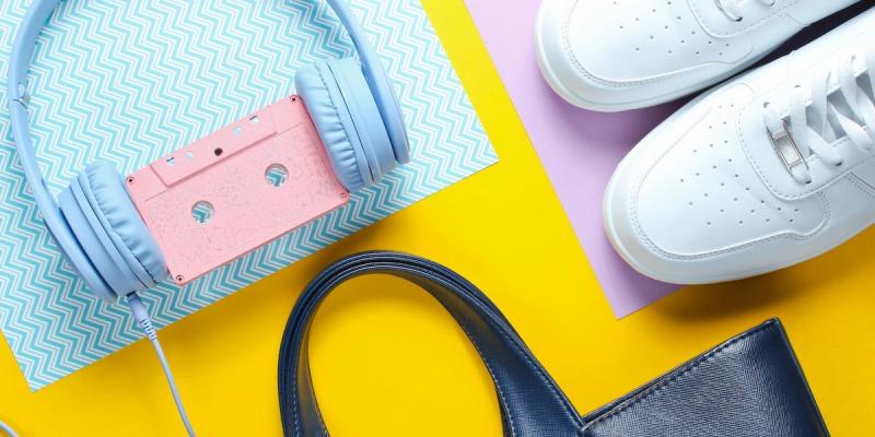 Flat-lay of blue headphones, pink cassette, white sneakers and navy leather bag on yellow background