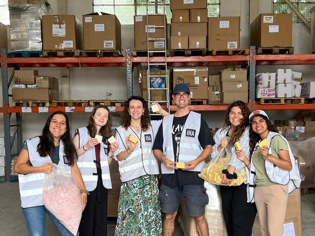 Six aid workers in safety vests smiling in a warehouse stocked with relief supply boxes