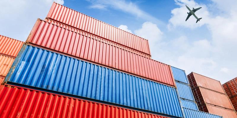 Stacked red, blue and orange shipping containers with an airplane flying overhead in a blue sky.