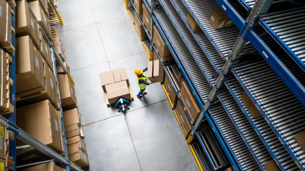 Aerial view of a warehouse worker in a hi-vis vest and hard hat moving boxes on a pallet jack between tall storage racks
