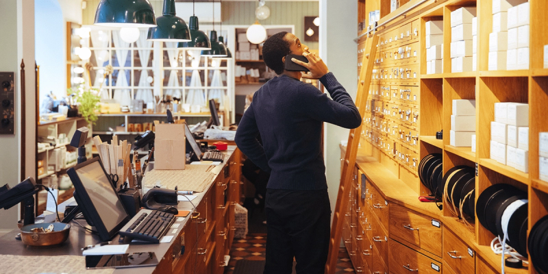 Man on a phone stands behind a shop counter beside wooden drawer shelving stocked with boxed inventory and a desktop monitor.