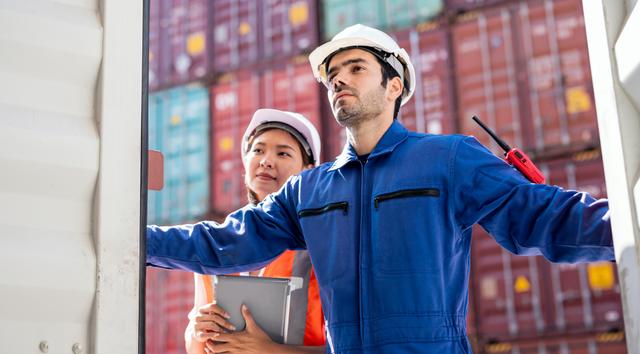 Two workers in hard hats and coveralls inspecting stacked shipping containers