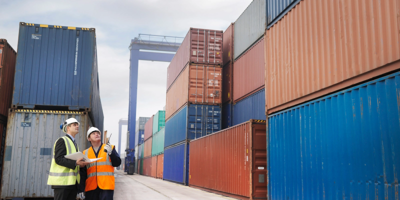 Two workers in hard hats and hi-vis vests stand in an aisle of stacked shipping containers at a port terminal