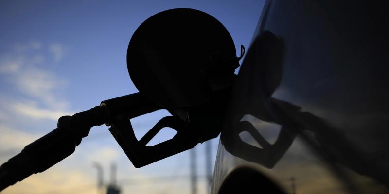 Silhouette of a fuel pump nozzle filling a vehicle against a dusk sky with smokestacks