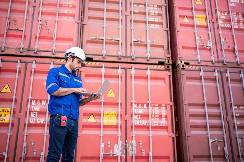 Worker in blue coveralls and white hard hat using a laptop in front of stacked red shipping containers