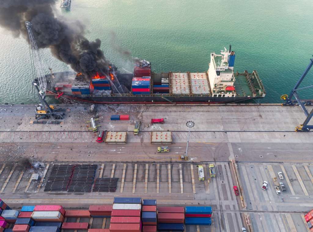 Aerial view of a container ship on fire at a port berth with a black smoke plume rising over the dock