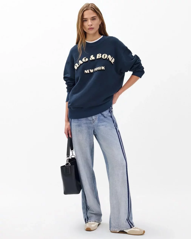Model wearing a Rag & Bone sweatshirt and jeans holding a handbag against a white studio backdrop