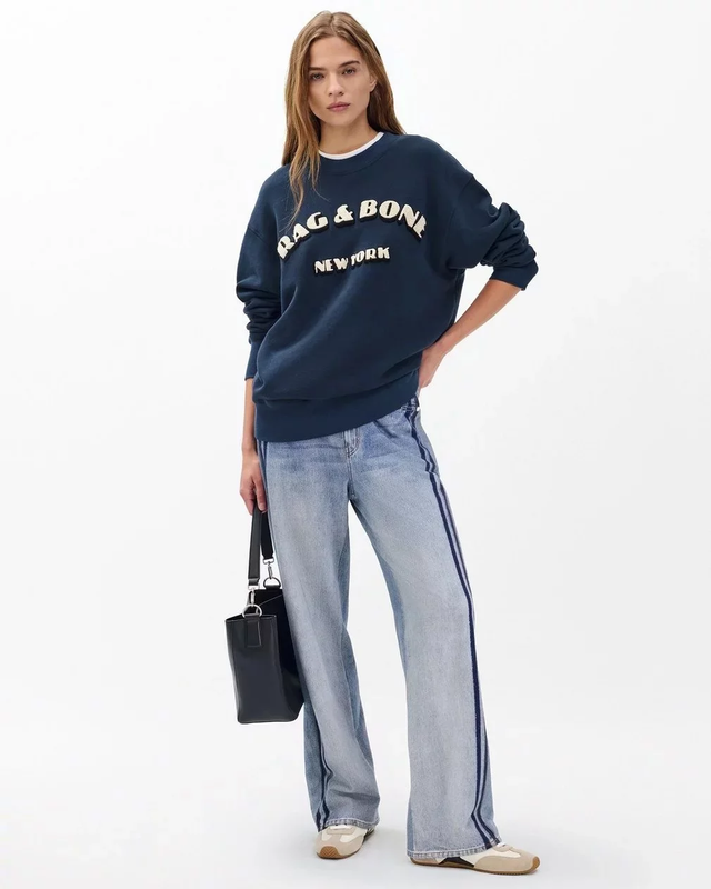 Model wearing a Rag & Bone sweatshirt and jeans holding a handbag against a white studio backdrop