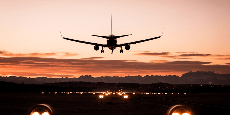 Silhouette of an airliner landing toward lit runway lights against an orange sunset sky