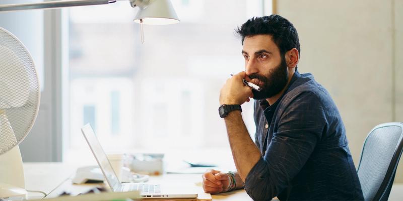 Bearded man in blue shirt at a desk gazing past his laptop, pen and phone in hand, with a fan nearby
