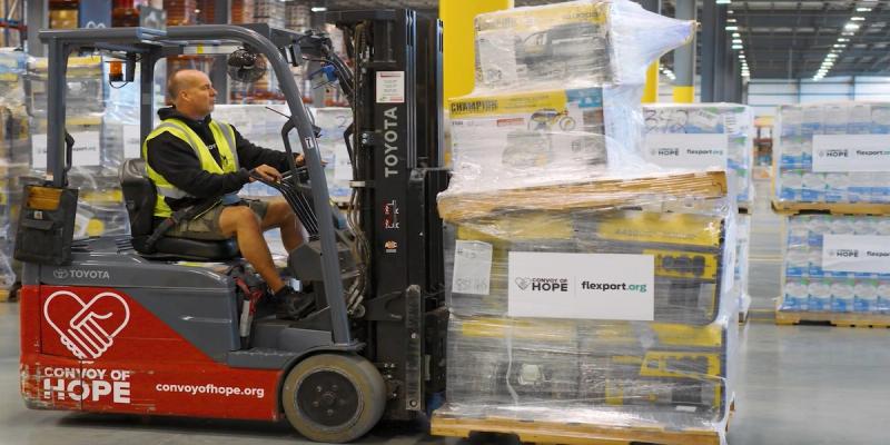 Worker drives a Toyota forklift branded Convoy of Hope moving shrink-wrapped pallets labeled flexport.org in a warehouse