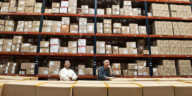 Two warehouse workers sitting amid tall racks of stacked cartons, evoking inventory financing and trade.