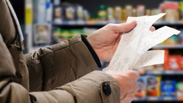A shopper in a winter coat holds a long paper grocery receipt while standing in a stocked supermarket aisle