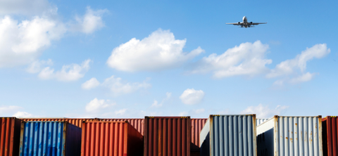 Photo of a passenger jet climbing over a row of stacked shipping containers against a blue partly cloudy sky.
