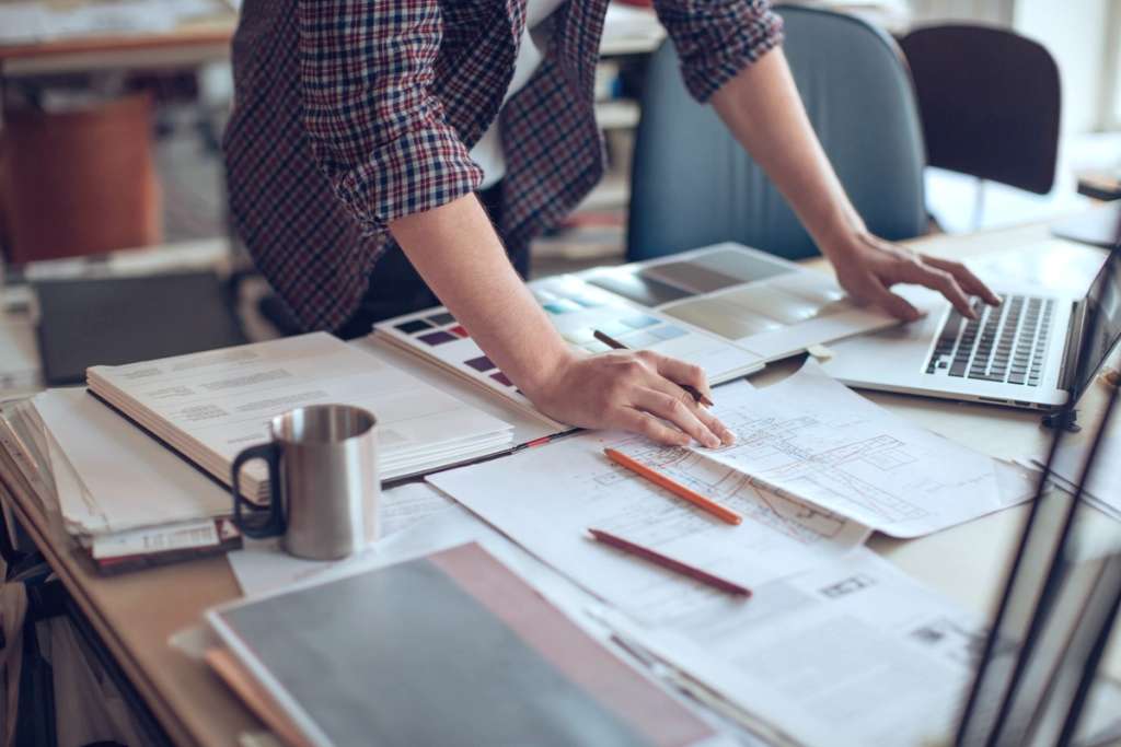 Person reviewing customs paperwork and blueprints at a desk beside a laptop