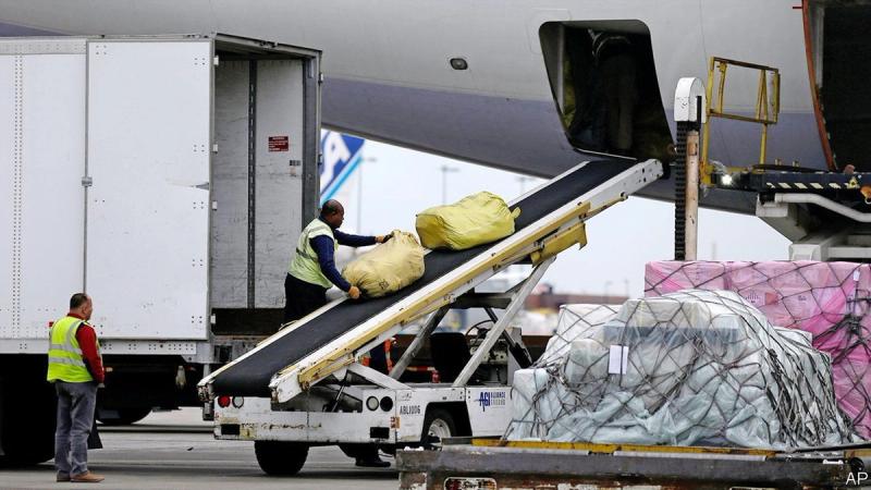 Workers in safety vests load cargo from a truck onto a conveyor into an aircraft on the tarmac, with wrapped freight pallets nearby