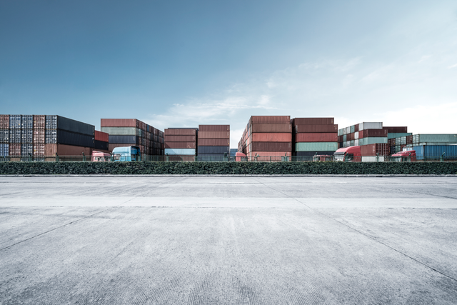 Rows of stacked shipping containers behind a wall beside an empty paved yard under a clear sky