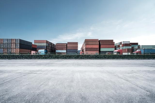 Rows of stacked shipping containers behind a wall beside an empty paved yard under a clear sky