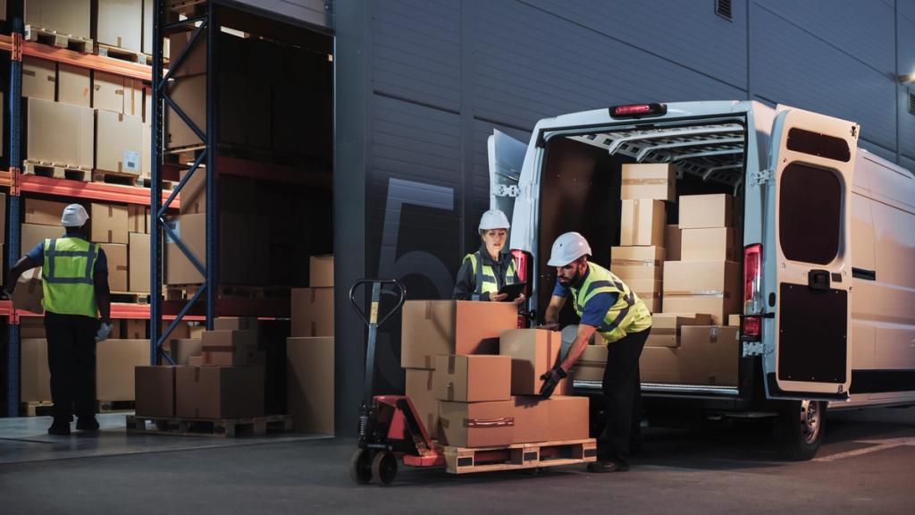 Two workers in hard hats and hi-vis vests loading boxes from a pallet jack into a delivery van at a dimly lit warehouse loading bay