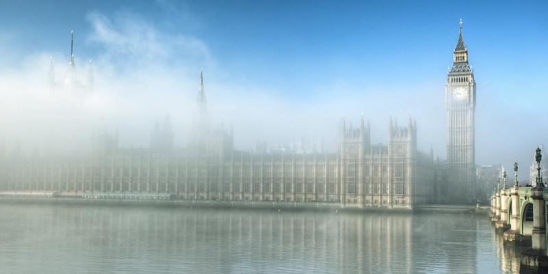 London's Houses of Parliament and Big Ben clock tower along the Thames, partly shrouded in fog under blue sky