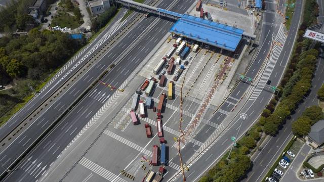 Aerial view of cargo trucks lined up at a sparsely occupied highway toll plaza with empty multilane roads