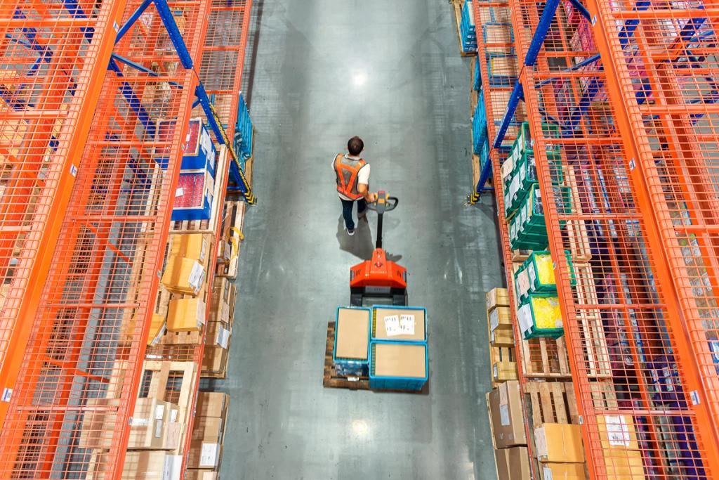 Overhead view of a warehouse worker on a powered pallet jack moving boxes down an aisle between tall orange storage racks