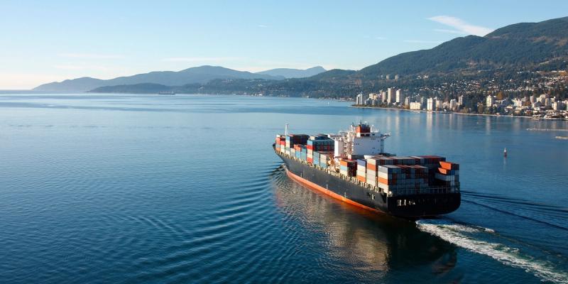 A container ship sails on calm blue water toward a coastline of forested hills and a waterfront city