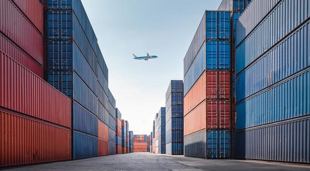 Rows of stacked shipping containers in a port yard with a passenger plane flying through the clear sky overhead
