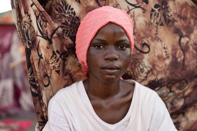Portrait of a young woman in a pink headscarf and white shirt seated against patterned fabric