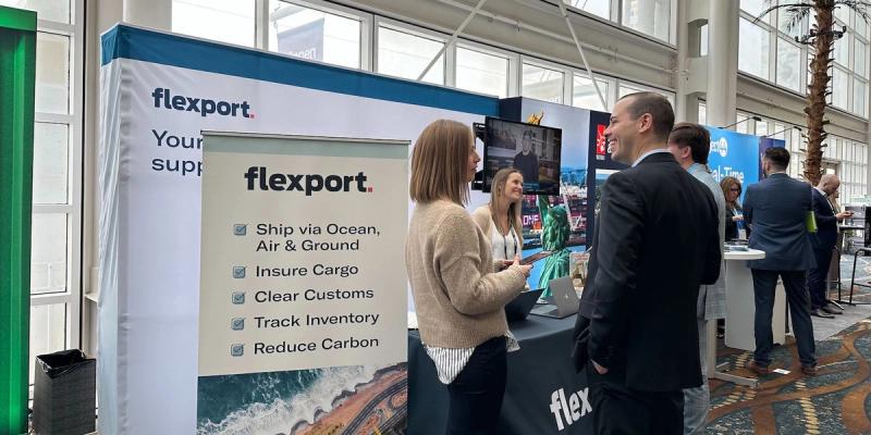 Flexport branded trade-show booth listing services as staff talk with attendees in a sunlit hall