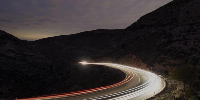 Long-exposure night shot of red and white car light trails winding along a curved mountain highway