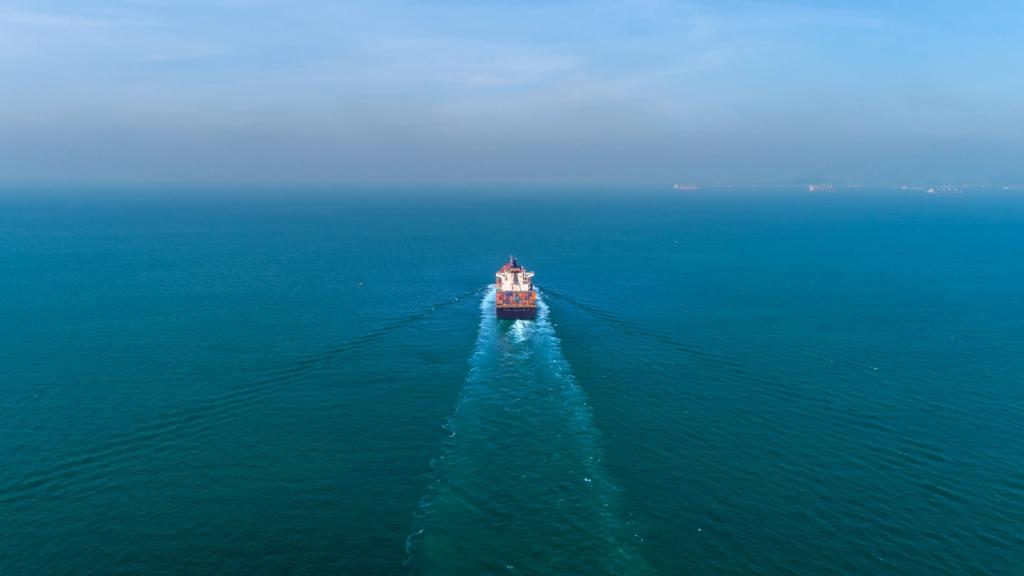 Aerial view of a container ship sailing alone across calm blue open ocean leaving a white wake, distant port cranes on horizon