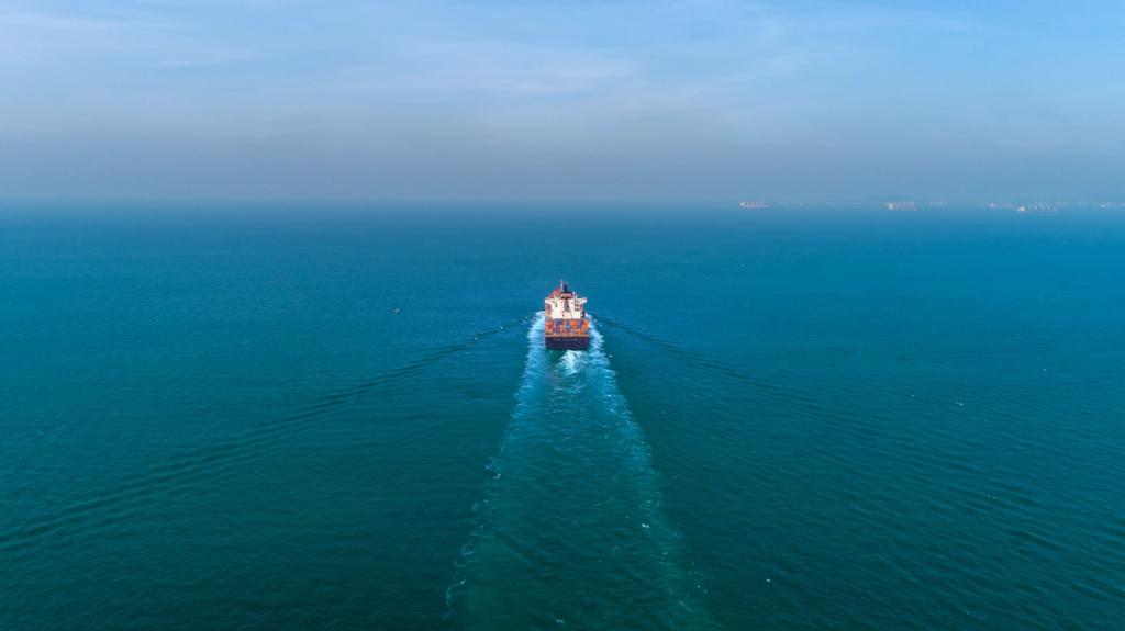 Aerial view of a container ship sailing alone across calm blue open ocean leaving a white wake, distant port cranes on horizon