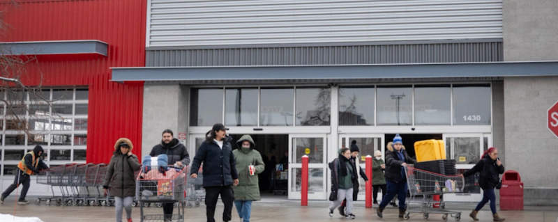 Shoppers in winter coats leave a big-box retail store, some pushing carts, on a cold overcast day
