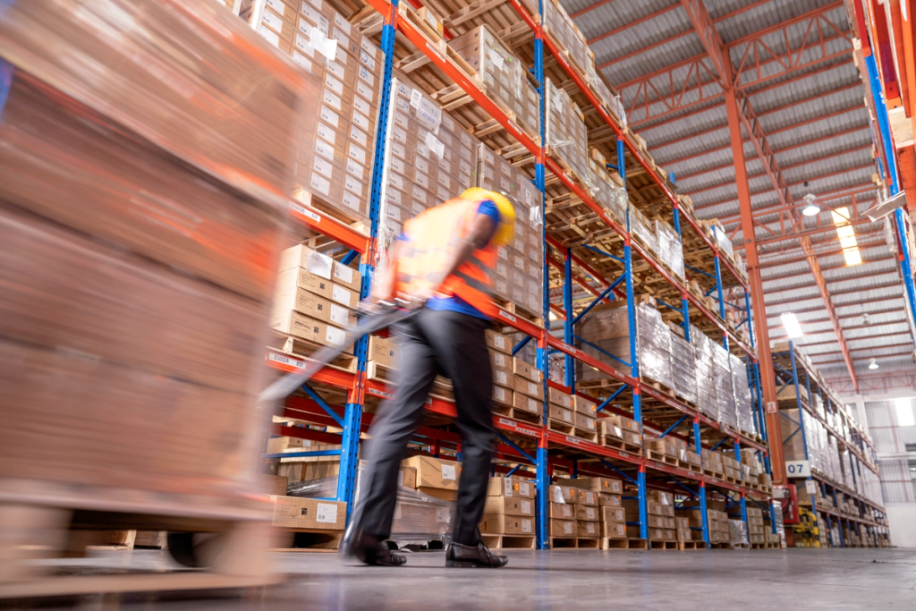 Worker in safety vest pulls a loaded pallet jack down an aisle of a high-rack warehouse