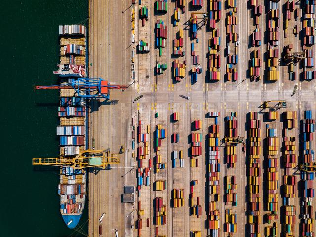 Aerial view of a container terminal with cranes loading a ship and color-blocked rows of stacked containers.