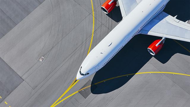 Aerial top-down view of a white passenger jet with red engines taxiing on a grey airport apron marked with yellow guide lines