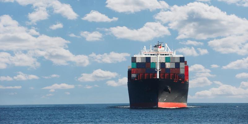 Container ship stacked with colorful containers sailing head-on across open water under a cloudy blue sky