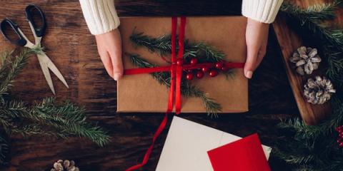 Overhead view of hands wrapping a kraft-paper gift with red ribbon, holly and pine sprigs on a wooden table.