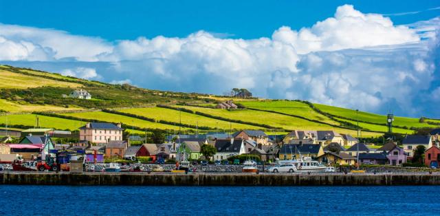 Colorful seaside Irish village with houses and a harbor below green patchwork hills