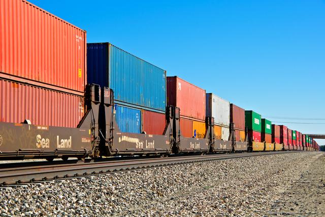 Line of railcars carrying stacked shipping containers, some marked Sea-Land, beside tracks under blue sky