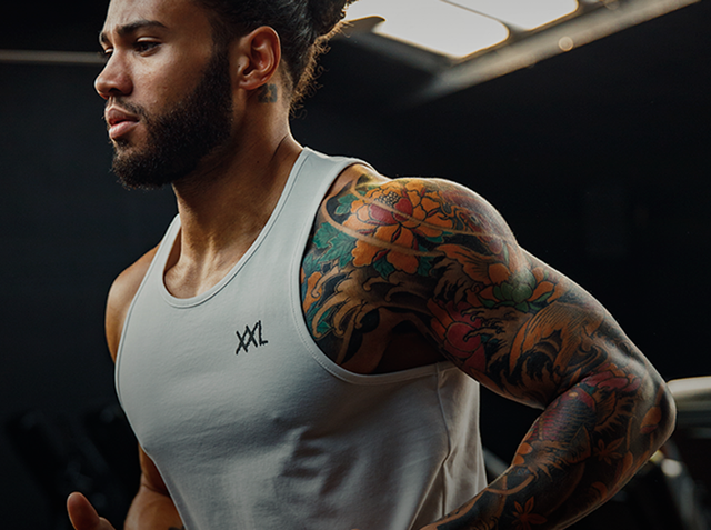 Athletic man in a white XXL Nutrition tank top with tattooed arm in a dark gym