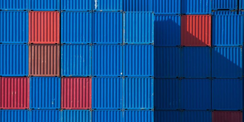 Wall of stacked shipping containers, mostly blue with scattered red ones, casting a diagonal shadow