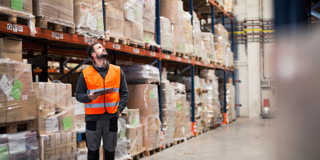 Warehouse worker in a hi-vis vest walking down an aisle of stocked shelves, representing fulfillment and logistics operations