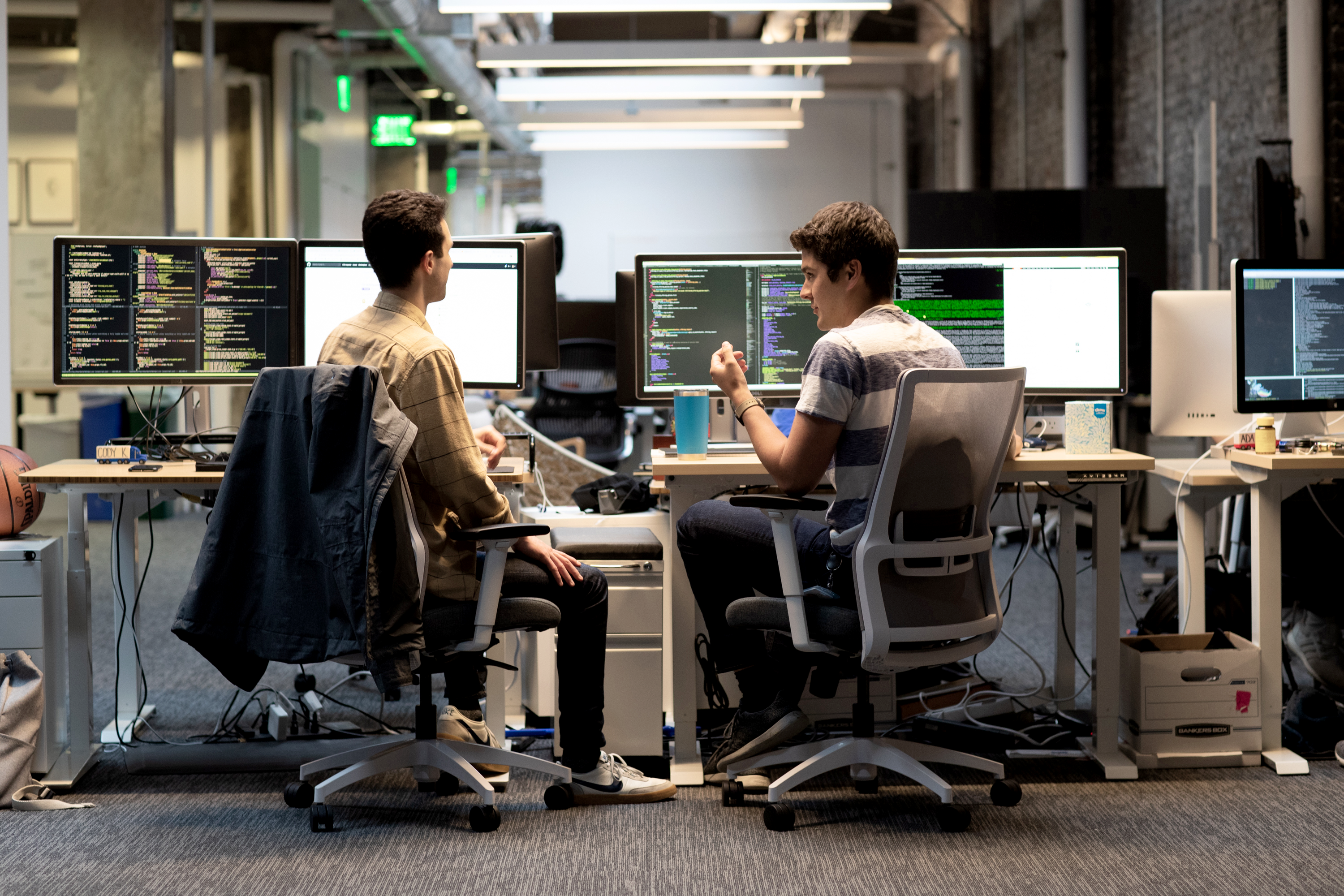 Two software developers working at desks with multiple monitors displaying code in a tech office.