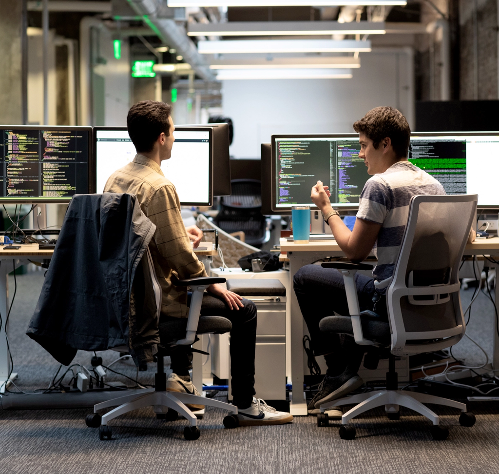 Two software developers working at desks with multiple monitors displaying code in a tech office.