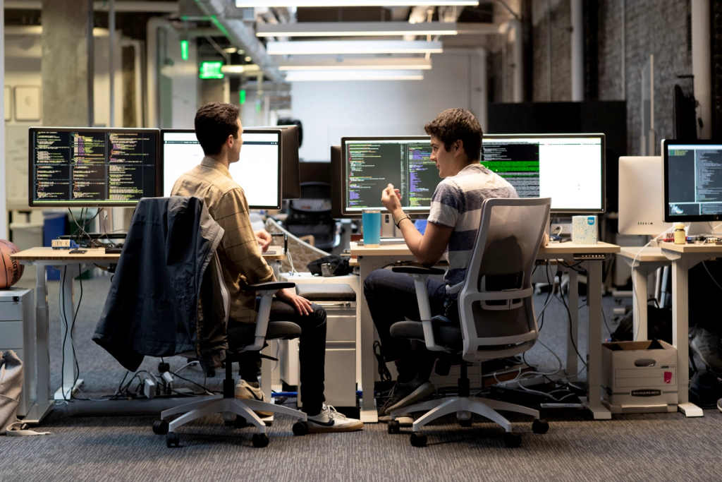 Two software developers working at desks with multiple monitors displaying code in a tech office.