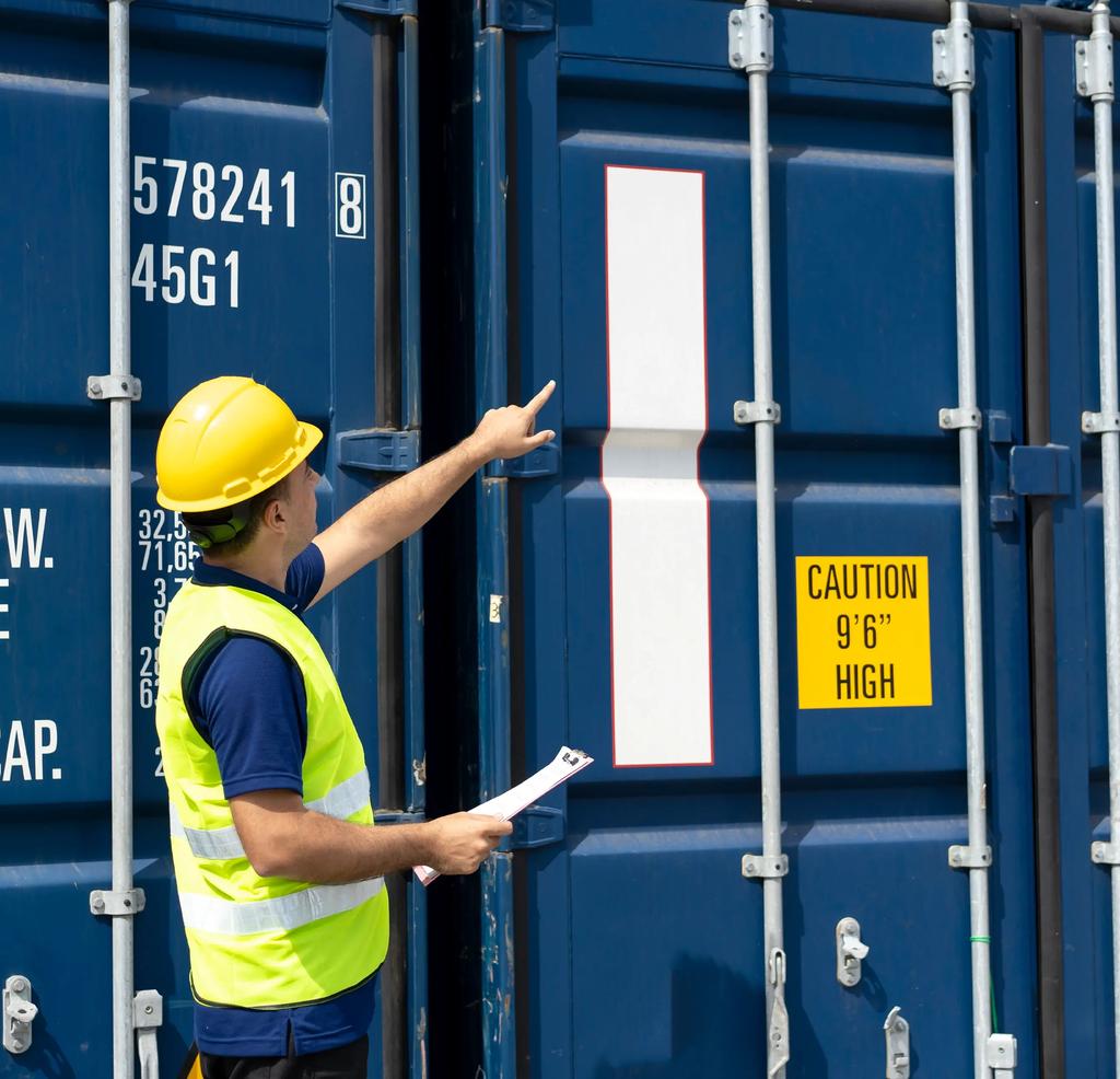 Photo of a worker in a hard hat and high-visibility vest holding a clipboard and pointing up at the markings on a blue shipping container.